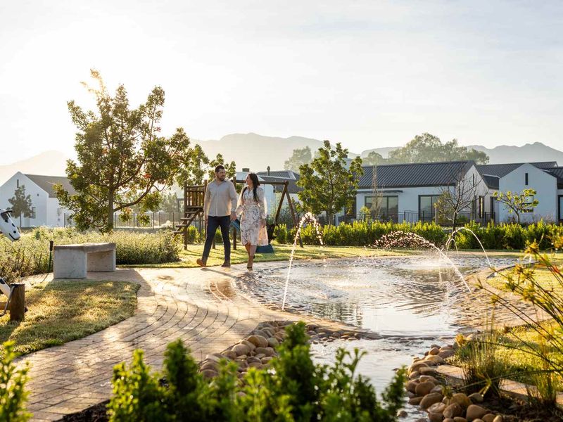 A beautifully landscaped residential garden featuring a central water feature, paved pathways, and a stone bench, set against a backdrop of modern homes.