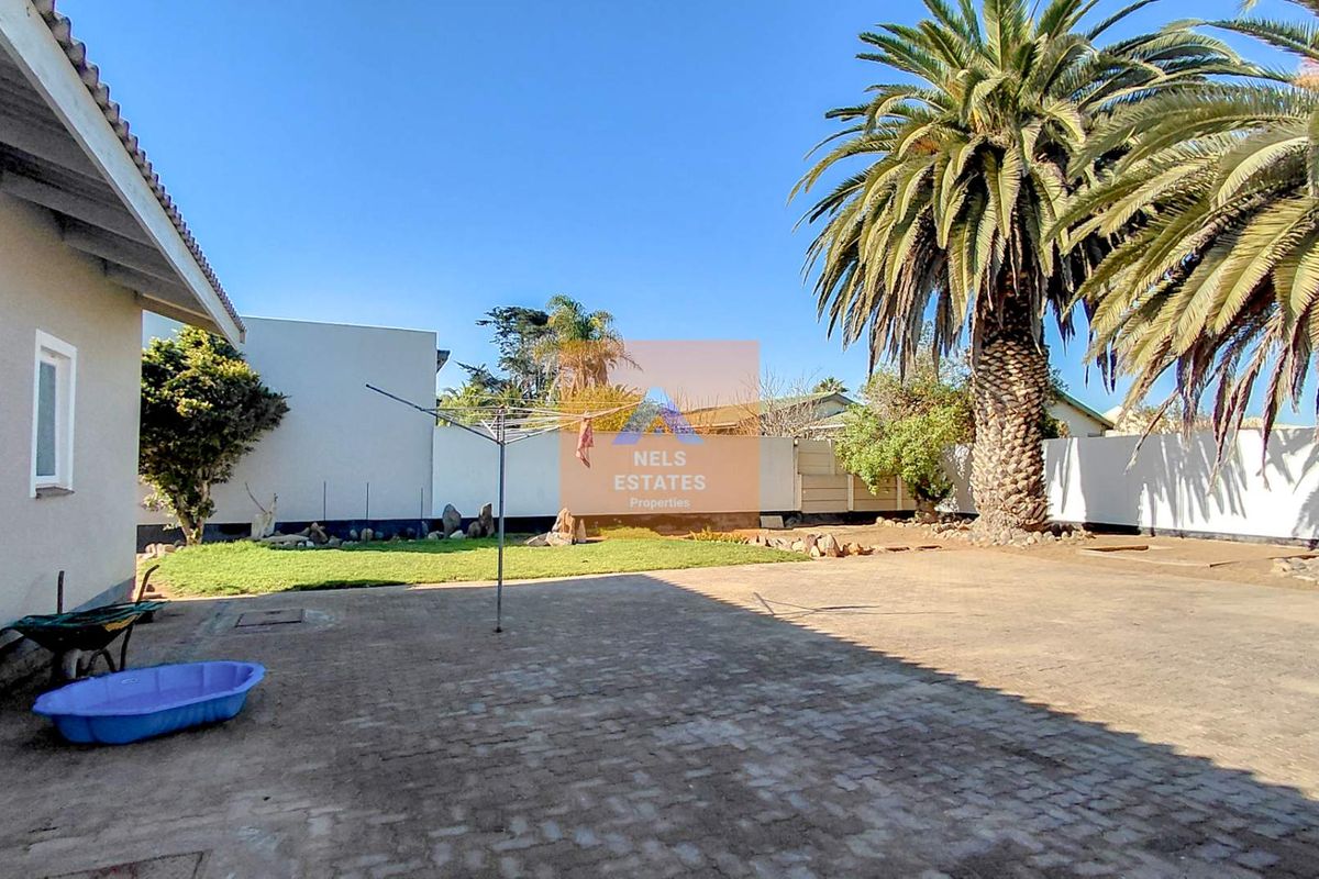 A spacious rear exterior area featuring a paved driveway, established palm tree, and a private garden enclosed by boundary walls.