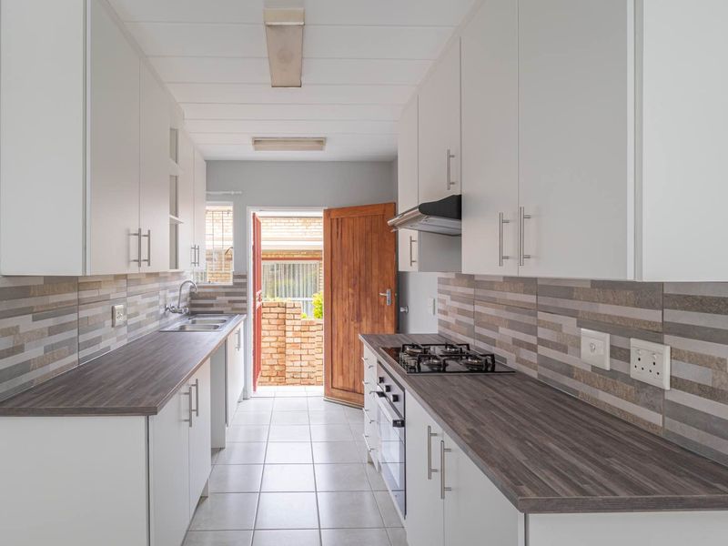 A clean, galley-style kitchen featuring white cabinetry, dark laminate countertops, a gas hob, and a tiled backsplash, leading to an exterior doorway.