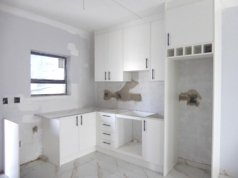 An unfinished kitchen space featuring white cabinetry, marble-look floor tiles, and dedicated fridge housing, currently awaiting final wall finishes and appliance installation.