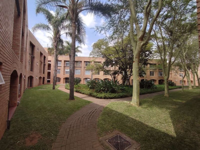 A well-maintained communal garden area featuring brick apartment facades, mature trees, and paved walkways within the residential complex.