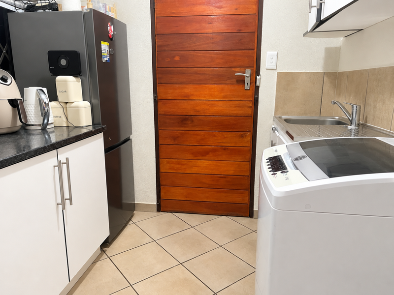 A functional laundry area featuring built-in white cabinetry, a stainless steel sink, tiled backsplash, and durable floor tiles with a solid wooden door.
