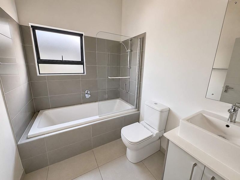 A modern, clean bathroom featuring a built-in bathtub with a glass shower screen, a contemporary vanity with basin, and neutral grey tiling.