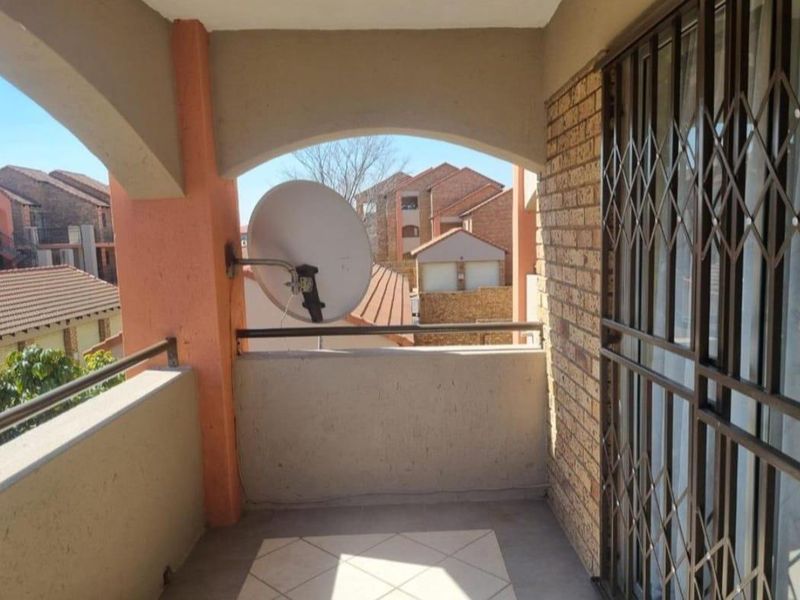 A covered balcony featuring tiled flooring, brick accents, and a metal security gate, offering an outdoor space with views of the surrounding complex.