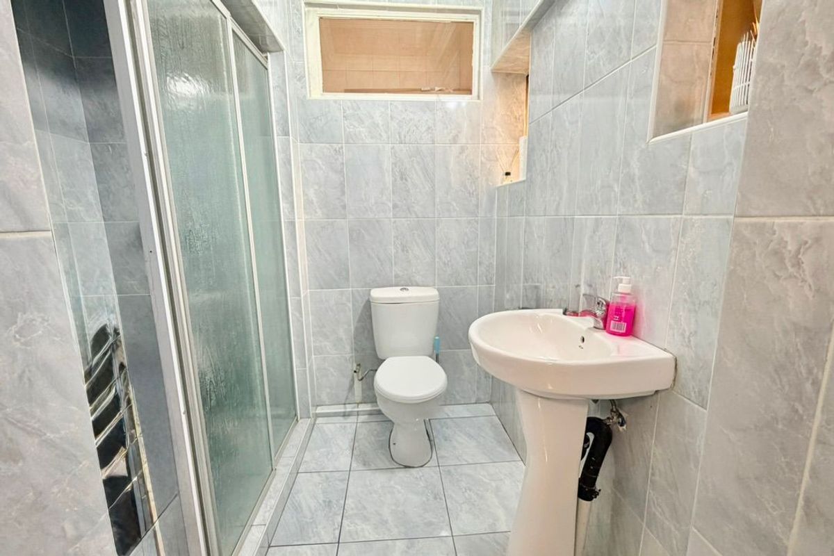 A compact bathroom featuring a shower enclosure, pedestal basin, and toilet, finished with light-toned wall and floor tiles.