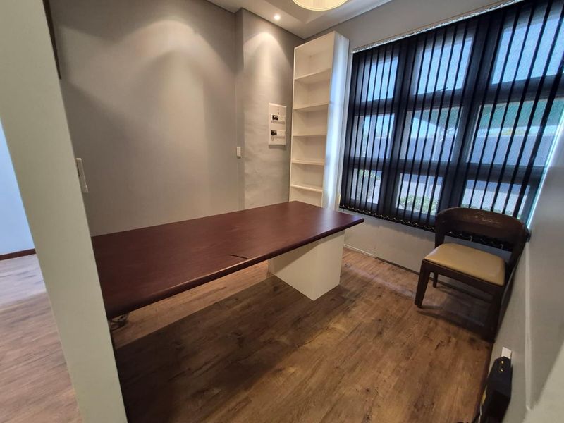 A functional office space featuring wood-look laminate flooring, built-in shelving, and large windows with vertical blinds, providing a professional work environment.