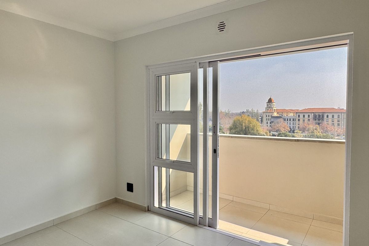 A bright, empty bedroom featuring neutral tiled flooring and a large sliding glass door that opens onto a private balcony with urban views.