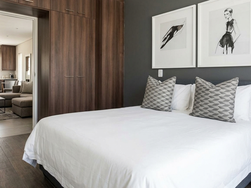 Modern bedroom featuring dark wood built-in wardrobes, tiled flooring, and recessed lighting, with an open doorway leading to an adjacent living area.