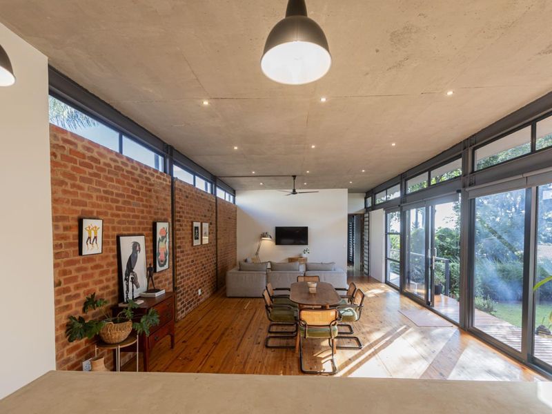 Spacious open-plan living and dining area featuring exposed brick walls, concrete ceilings, timber floors, and expansive sliding glass doors opening to the outdoors.