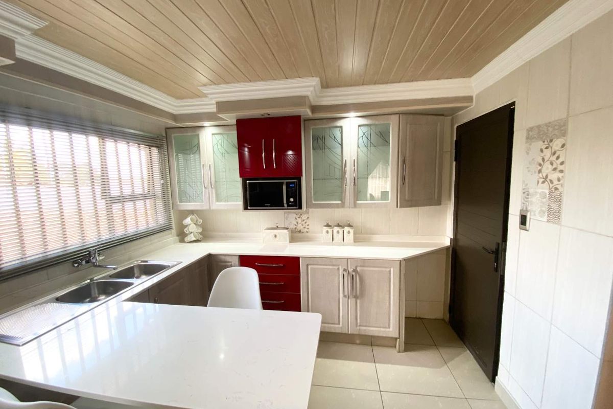 A well-maintained kitchen featuring light-toned cabinetry with contrasting red accents, stone countertops, a double sink, and a modern tiled ceiling.