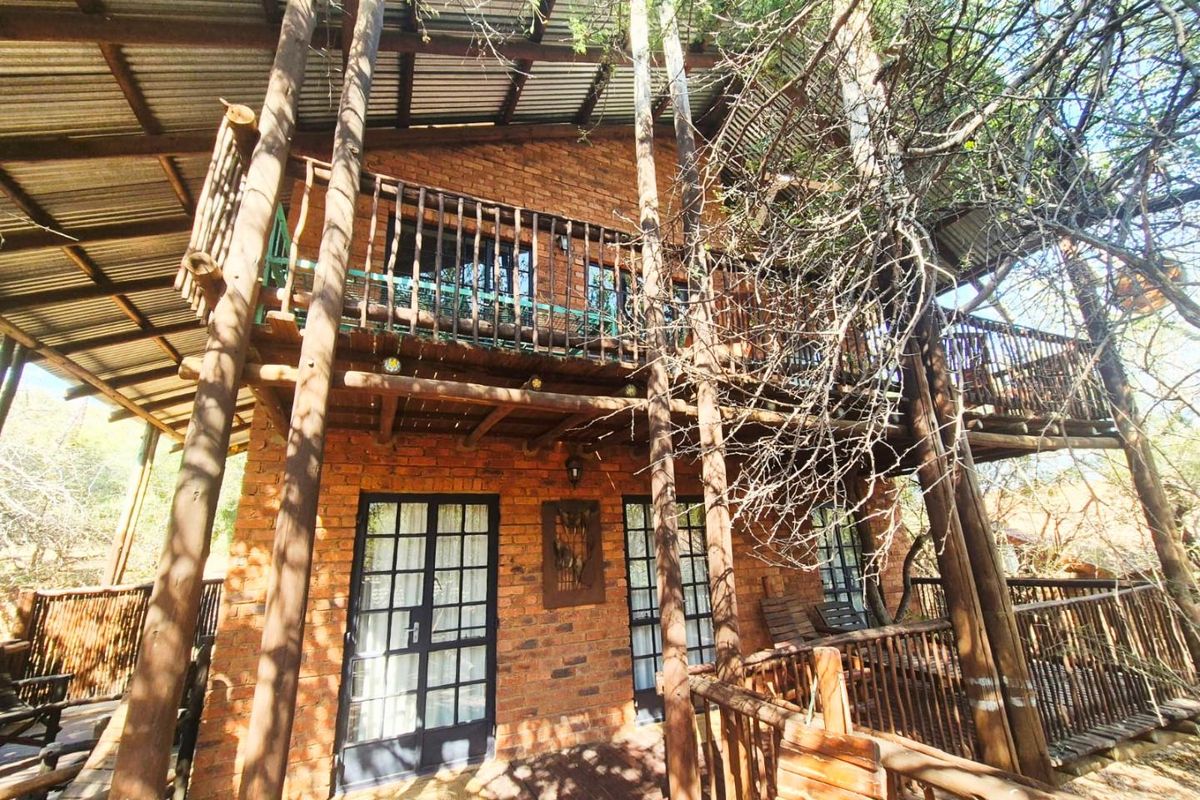 A rustic two-story brick house featuring timber balconies, wooden support pillars, and French doors, set within a natural wooded environment.