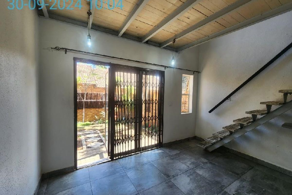 An open-plan living area featuring tiled floors, exposed timber ceiling beams, and a staircase, with direct access to the rear garden through a security gate.