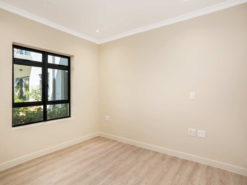 A bright, empty bedroom featuring modern laminate flooring, neutral walls, recessed lighting, and a built-in cupboard with a black-framed window.