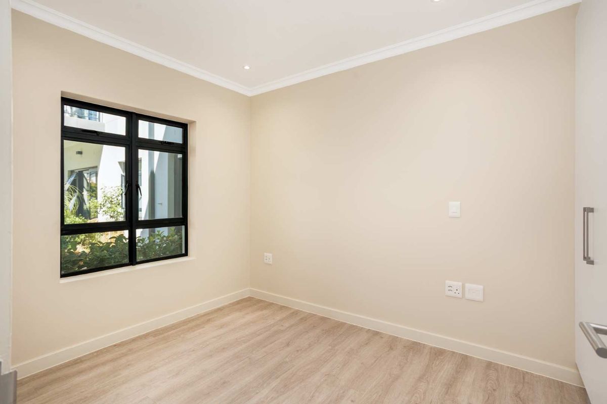A bright, empty bedroom featuring modern laminate flooring, neutral walls, recessed lighting, and a built-in cupboard with a black-framed window.