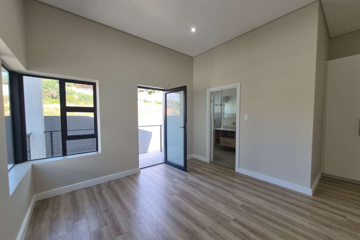 A bright, modern bedroom featuring wood-look flooring, recessed ceiling lighting, large windows, and direct access to an outdoor balcony and en-suite bathroom.