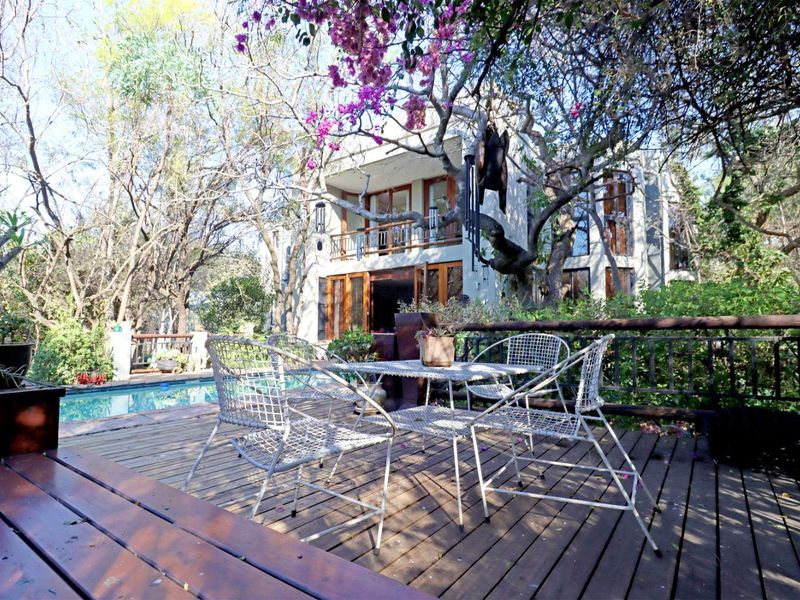 A wooden deck area featuring a swimming pool and views of the multi-story residence, surrounded by mature trees and garden foliage.