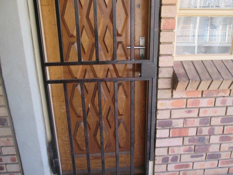 Exterior view of the apartment entrance featuring a solid wooden door protected by a black metal security gate and brick exterior wall.