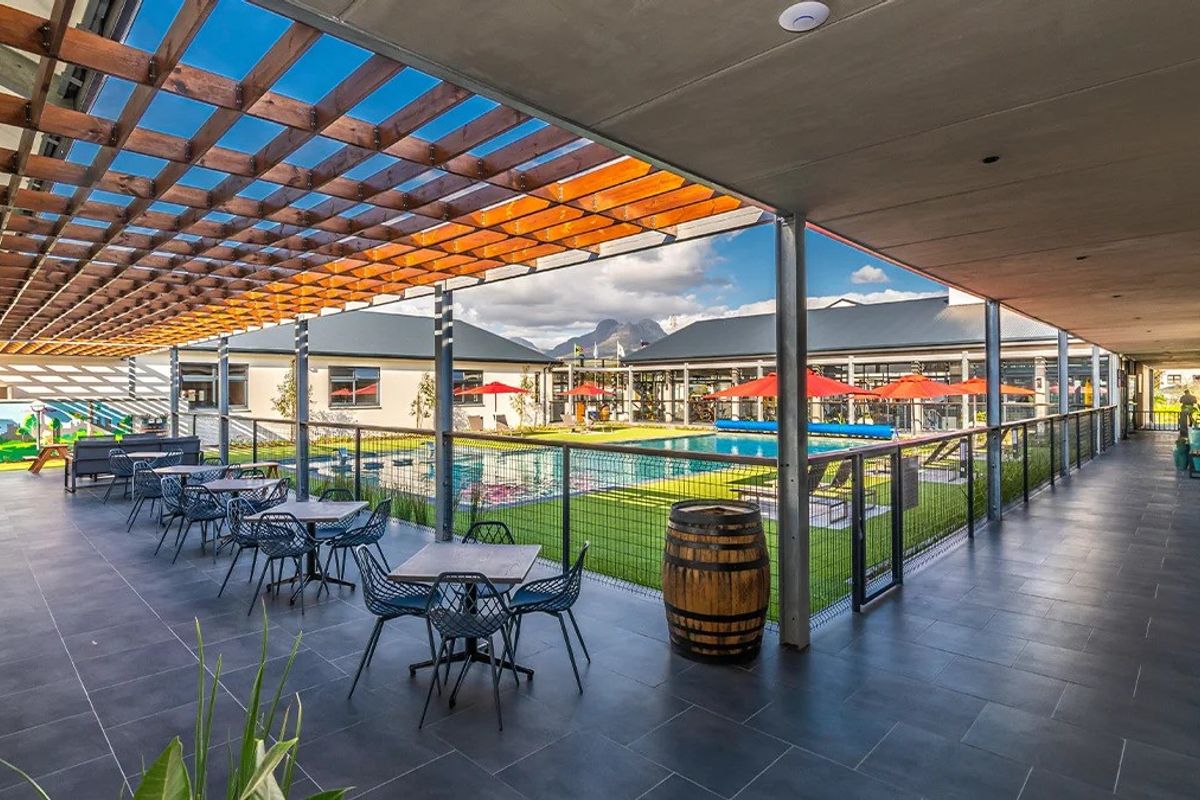 A spacious covered patio area overlooking a well-maintained swimming pool and landscaped garden, featuring modern tiled flooring and a timber pergola structure.