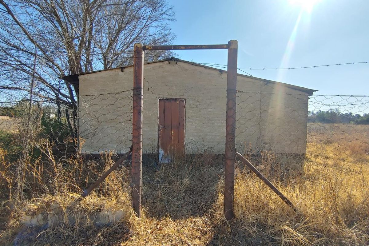 A weathered outbuilding on a small holding, featuring a single door, cracked masonry walls, and a rusted metal gate frame in a rural setting.