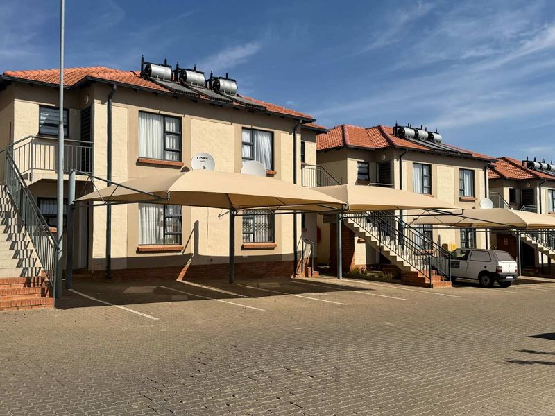 Exterior view of a multi-story apartment complex featuring paved parking, shade cloth carports, external staircases, and roof-mounted solar water heating systems.