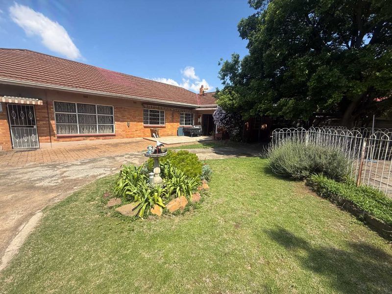 Front exterior view of a single-story brick house featuring a tiled roof, paved driveway, and established garden area with security fencing.