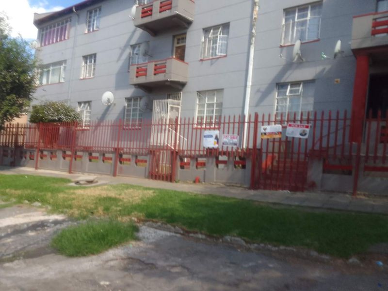 Exterior view of a multi-story apartment building featuring steel security fencing, balconies, and security gates at the entrance.