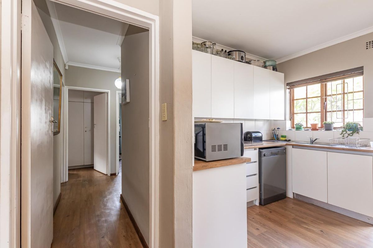 A functional kitchen featuring white cabinetry, wood-look flooring, and a window overlooking the exterior, adjacent to a hallway leading to other rooms.