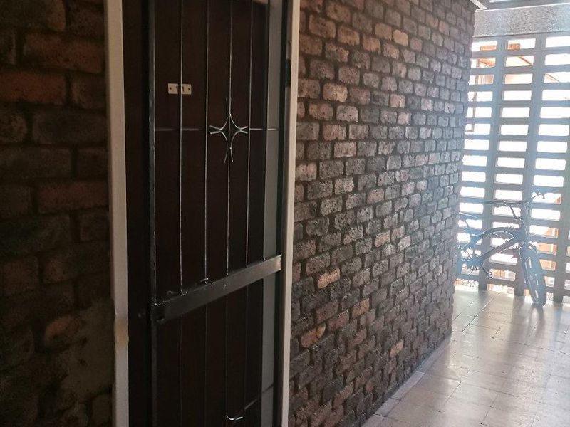 A narrow hallway featuring exposed brick walls, tiled flooring, and a security gate, leading to an outdoor breeze block screen area.
