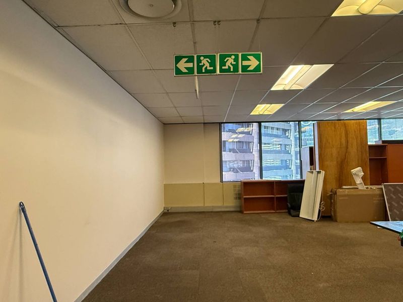 An open-plan office space featuring a suspended ceiling with recessed lighting, large windows, and carpeted flooring, currently in need of refurbishment.