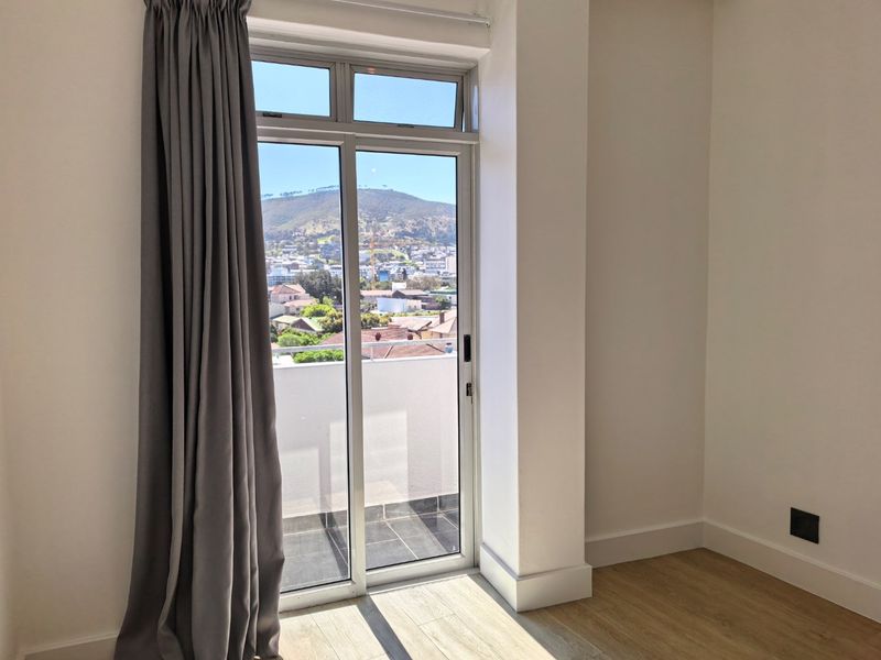 A bright, empty bedroom featuring modern laminate flooring, clean white walls, and a sliding door providing access to a balcony with scenic mountain views.