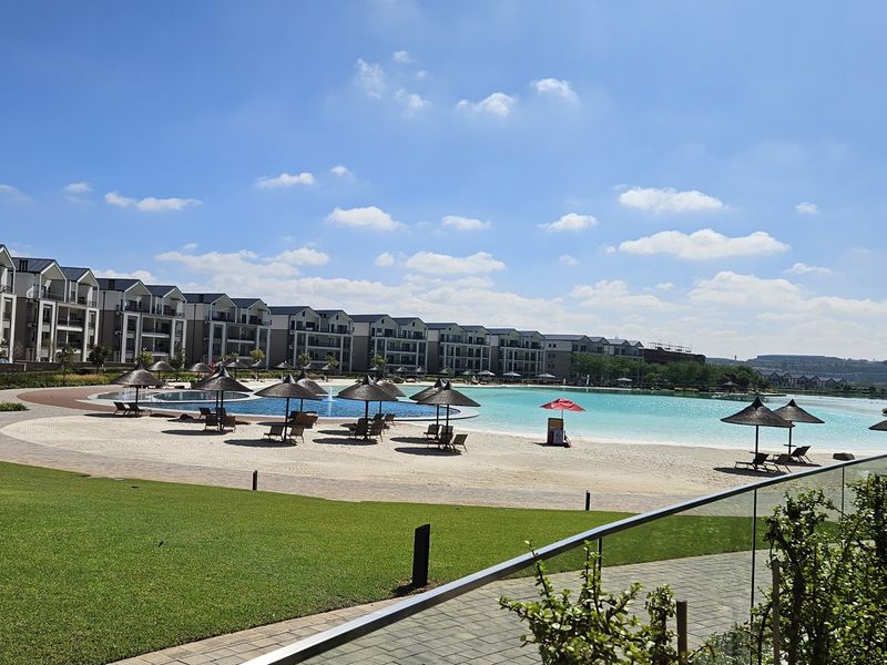 A well-maintained communal pool area featuring a large swimming pool, sandy beach area, and landscaped lawns within a modern apartment complex.