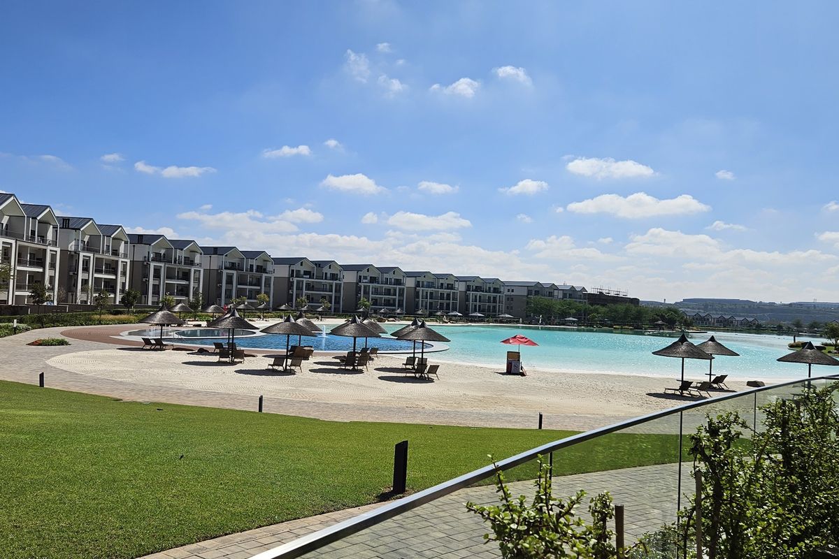 A well-maintained communal pool area featuring a large swimming pool, sandy beach area, and landscaped lawns within a modern apartment complex.