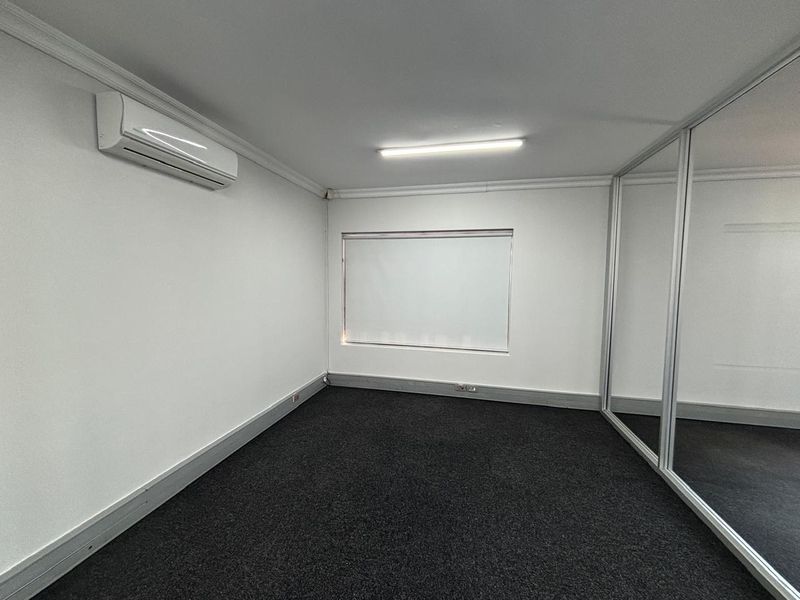 A clean, empty office space featuring neutral walls, dark carpeting, a split-system air conditioner, and a modern glass partition wall.