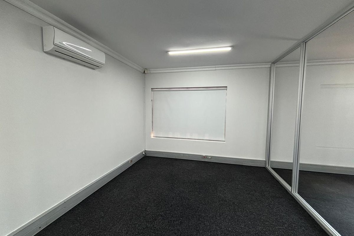 A clean, empty office space featuring neutral walls, dark carpeting, a split-system air conditioner, and a modern glass partition wall.