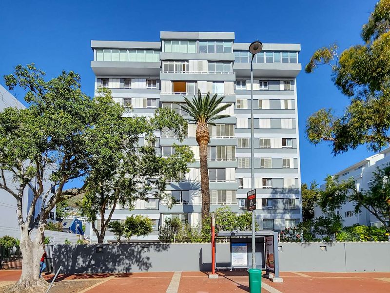 A clear street-level view of the front facade of a multi-story apartment building featuring balconies and established surrounding trees.