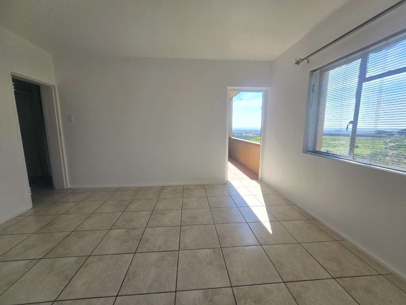 An empty living room featuring neutral tiled flooring, large windows with venetian blinds, and direct access to an outdoor balcony area.