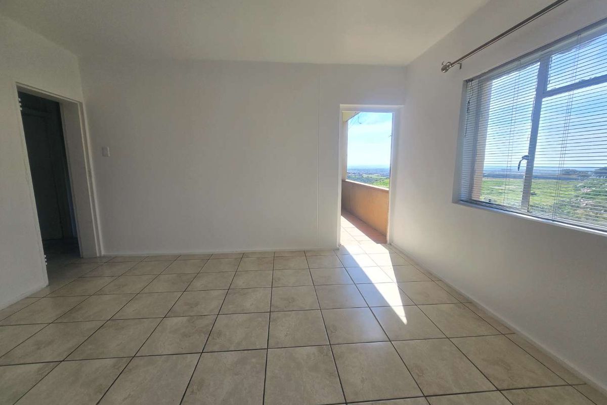 An empty living room featuring neutral tiled flooring, large windows with venetian blinds, and direct access to an outdoor balcony area.