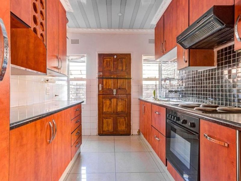 A functional galley-style kitchen featuring wood-finish cabinetry, dark tiled backsplash, built-in oven and hob, and tiled flooring.