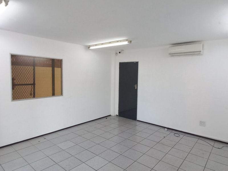 A clean, empty industrial office space featuring tiled flooring, fluorescent strip lighting, a wall-mounted air conditioning unit, and an internal viewing window.