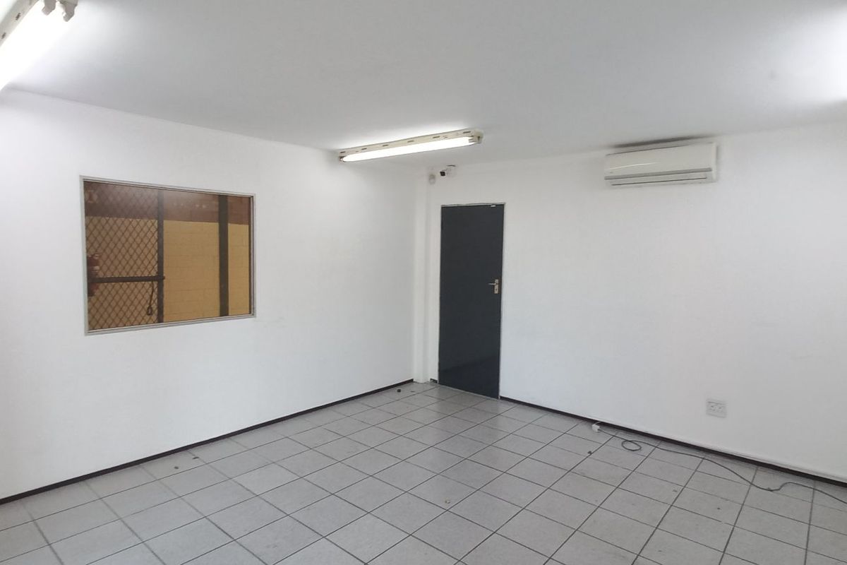 A clean, empty industrial office space featuring tiled flooring, fluorescent strip lighting, a wall-mounted air conditioning unit, and an internal viewing window.