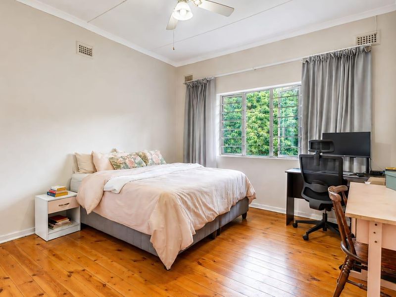 A well-lit bedroom featuring polished timber flooring, a ceiling fan, and large windows overlooking greenery, providing a comfortable and functional living space.