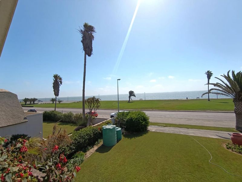 A scenic elevated view overlooking a coastal road, green lawn, and the ocean, featuring well-maintained garden areas and exterior water storage tanks.