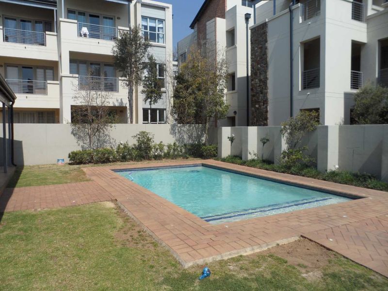 A communal outdoor pool area featuring a paved surround, landscaped garden, and boundary wall within a modern apartment complex.