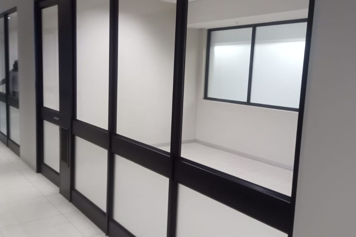 A bright, modern office hallway featuring glass partition walls, aluminum framing, and clean tiled flooring, providing a professional and open workspace environment.