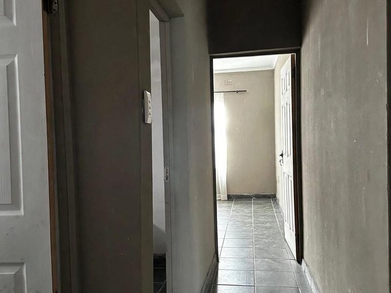 A narrow hallway featuring dark tiled flooring and unfinished wall surfaces, leading to an adjacent room with natural light.