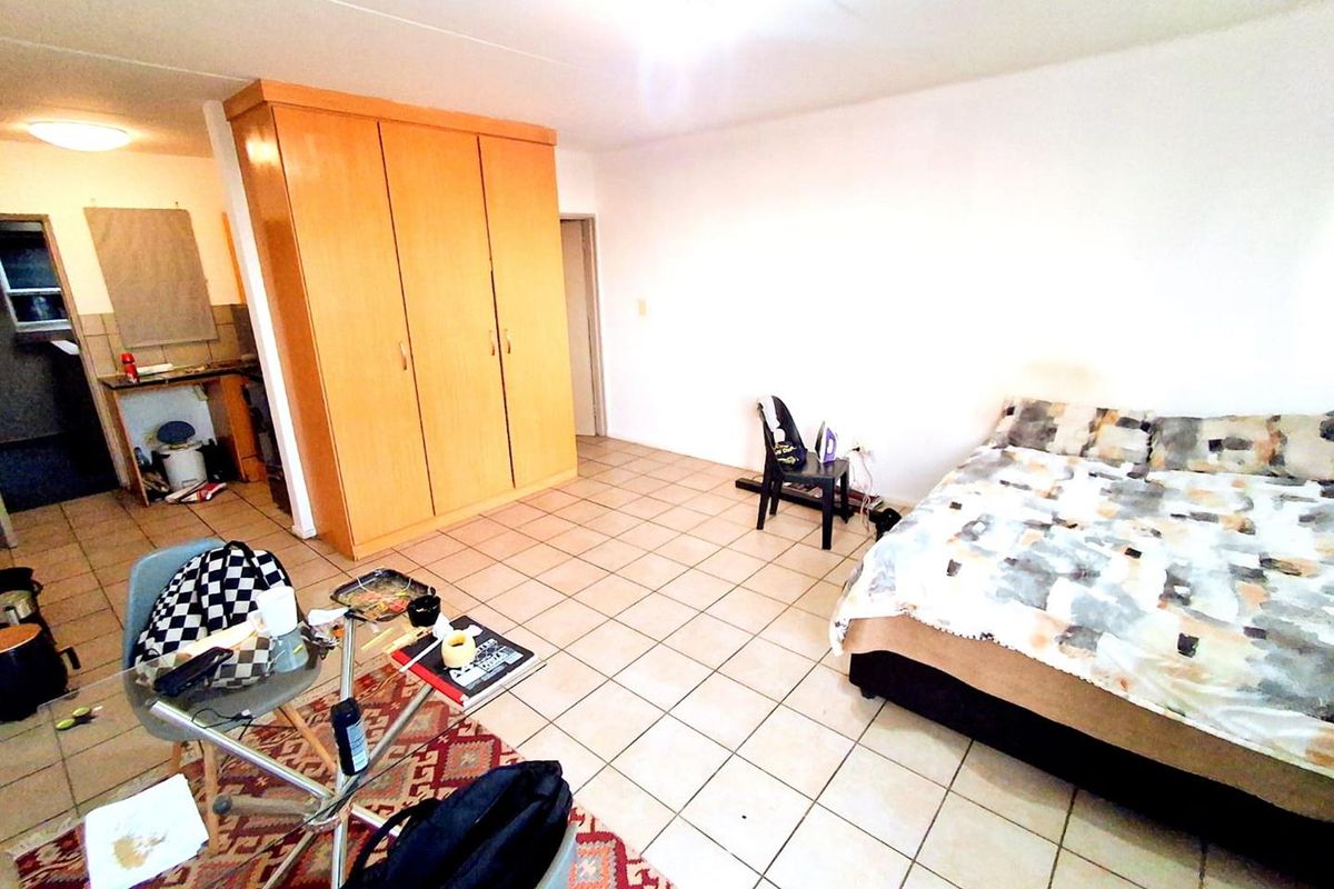 A bedroom featuring built-in wooden cupboards and tiled flooring, with an adjacent small kitchenette area visible in the background.