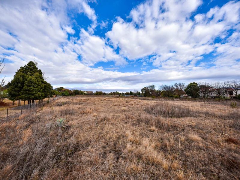 A wide-angle view of a vacant residential plot featuring dry grass and perimeter wire fencing under a bright, cloudy sky.