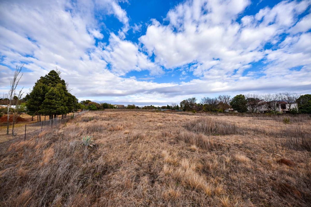 A wide-angle view of a vacant residential plot featuring dry grass and perimeter wire fencing under a bright, cloudy sky.