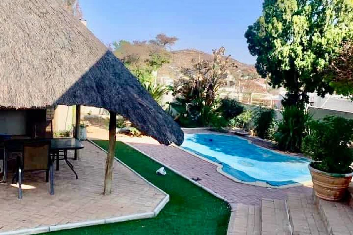 Outdoor entertainment area featuring a thatched lapa, paved patio, artificial lawn, and a swimming pool with a scenic hillside backdrop.
