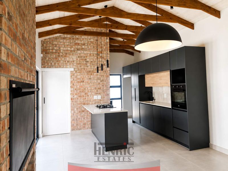 Modern kitchen featuring exposed timber trusses, brick feature wall, sleek dark cabinetry, stone countertops, gas hob, and a built-in fireplace.